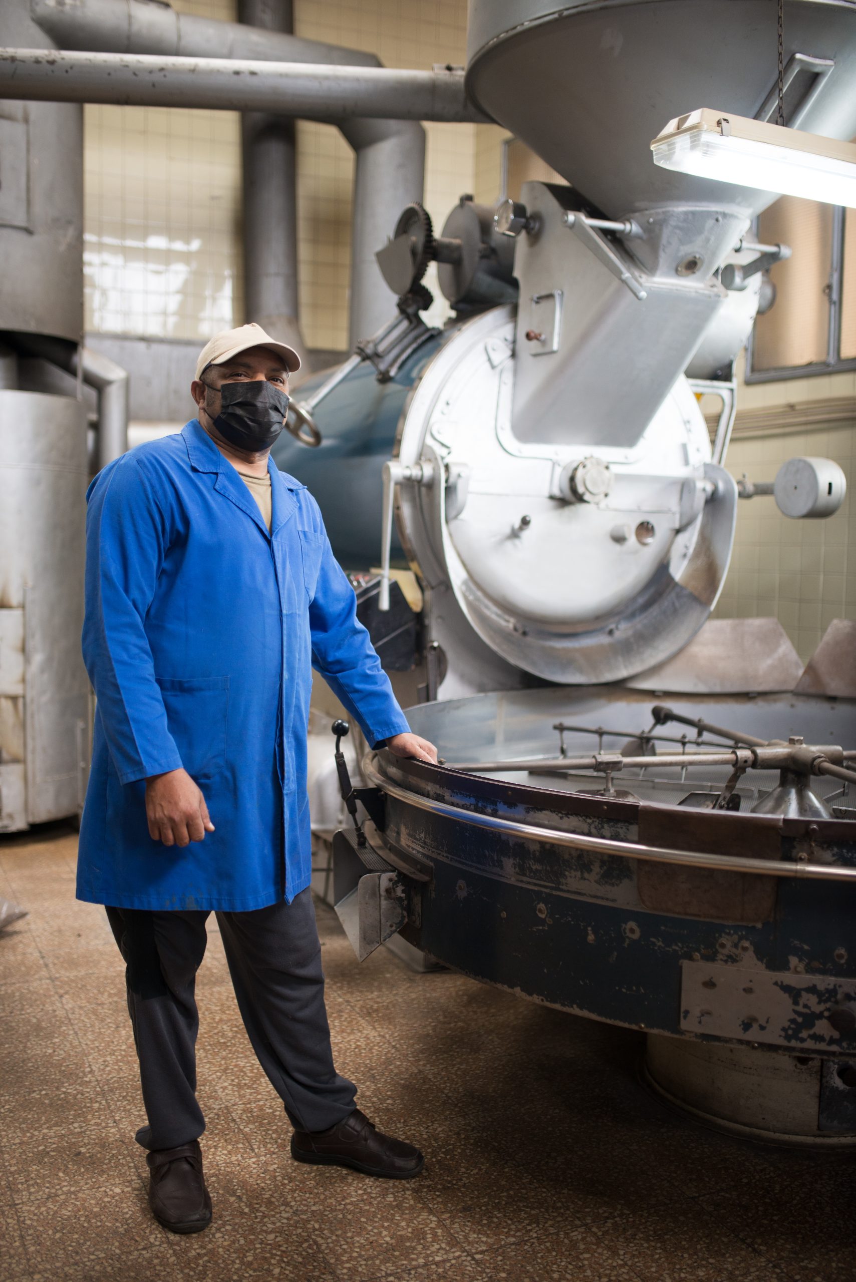 Mid adult coffee roaster standing next to coffee roasting machin Mid adult coffee roaster standing next to coffee roasting machine. Interior of coffee production workshop with set up roasting equipment. Person in uniform and mask. Production, food concept
